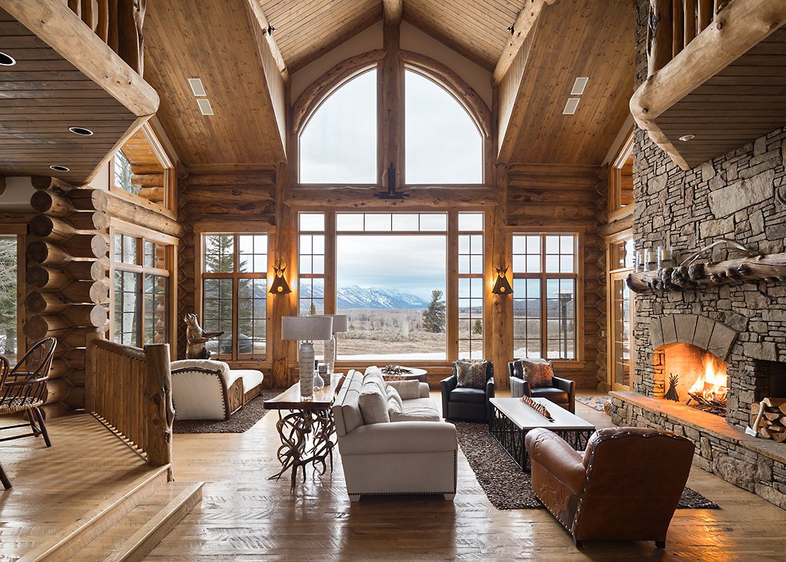 A luxurious and spacious rustic living room with a large stone fireplace, comfortable seating, and an expansive window view of snow-capped mountains.