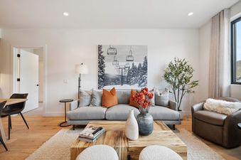 A bright and modern living room at Frontgate 226 featuring a gray sofa with orange and gray pillows, a unique wooden coffee table, a black-and-white ski lift art piece, a green plant, and a brown leather armchair with a white faux fur throw.