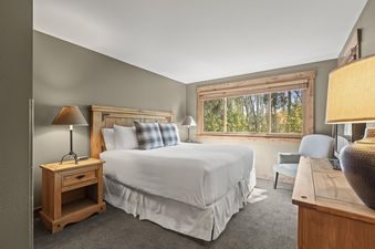A cozy bedroom with a king-sized bed, rustic wooden furniture, and a large window offering a view of green trees.