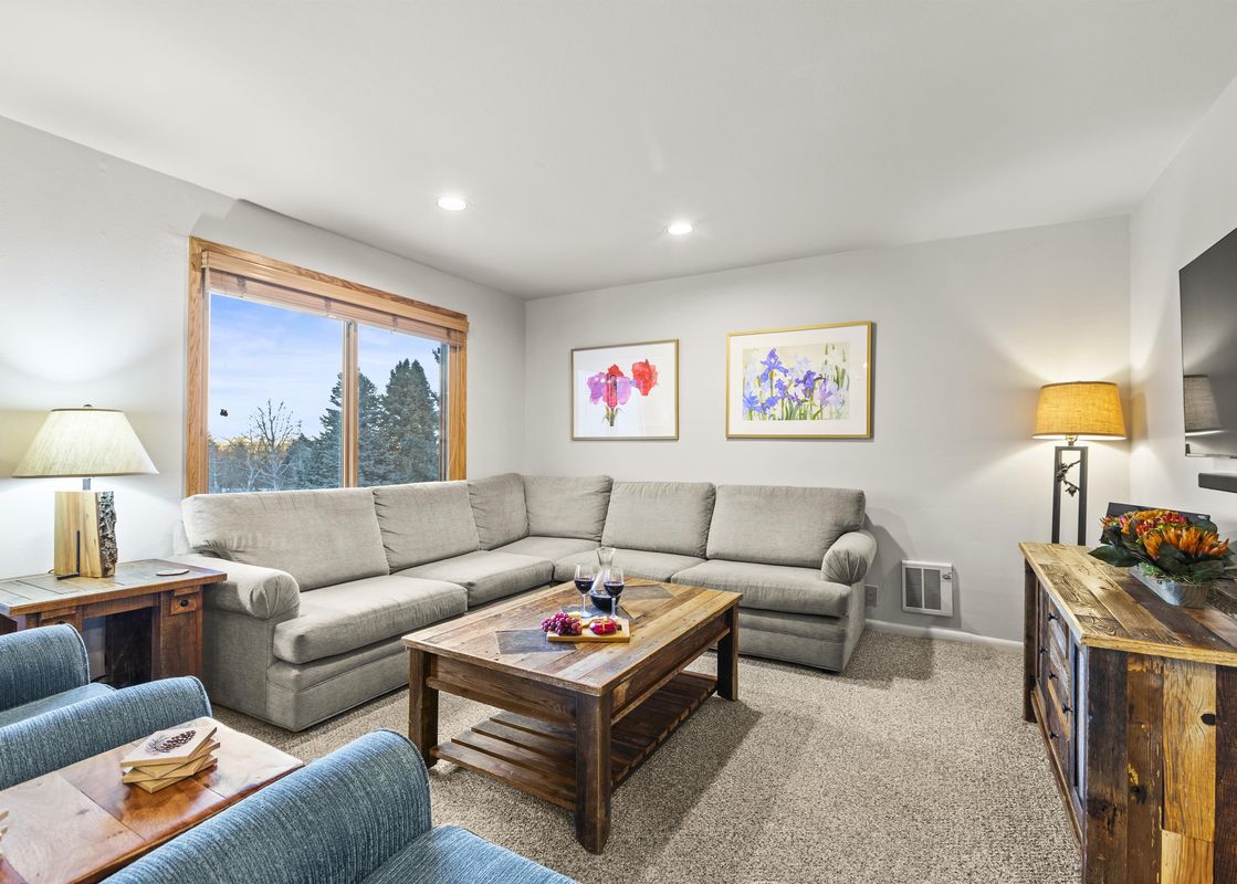 A cozy and inviting lounge area at Snow Flower 41 Park City, featuring a large L-shaped sofa, a rustic wooden coffee table with wine and snacks, and framed floral artwork on the walls.