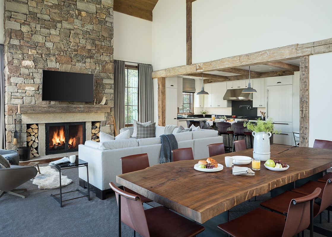 A spacious and luxurious great room in Fish Creek Lodge 11 features an open-concept living, dining, and kitchen area, centered around a grand stone fireplace with a roaring fire and a large wooden dining table set for a meal.
