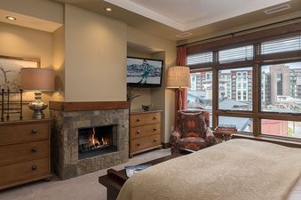 A luxurious bedroom at Longs Peak One Steamboat Place 609 featuring a fireplace, a mounted TV, a large window with a view of the town, a comfortable bed, and elegant furnishings.