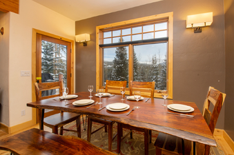 A formal dining area with a rustic wooden table set with plates and glasses, bathed in warm lighting. A large window offers a view of snow-covered pine trees and distant mountains.