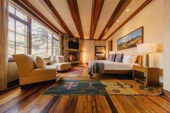 A cozy bedroom at Mount Wilson Horizons at See Forever features a large bed, stone fireplace, two armchairs, and views of pine trees through large windows.