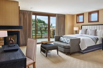 A luxurious bedroom at the St Regis 2 Bedroom Snow Park features a king-sized bed, a fireplace, and a large balcony overlooking a lush, treed landscape.