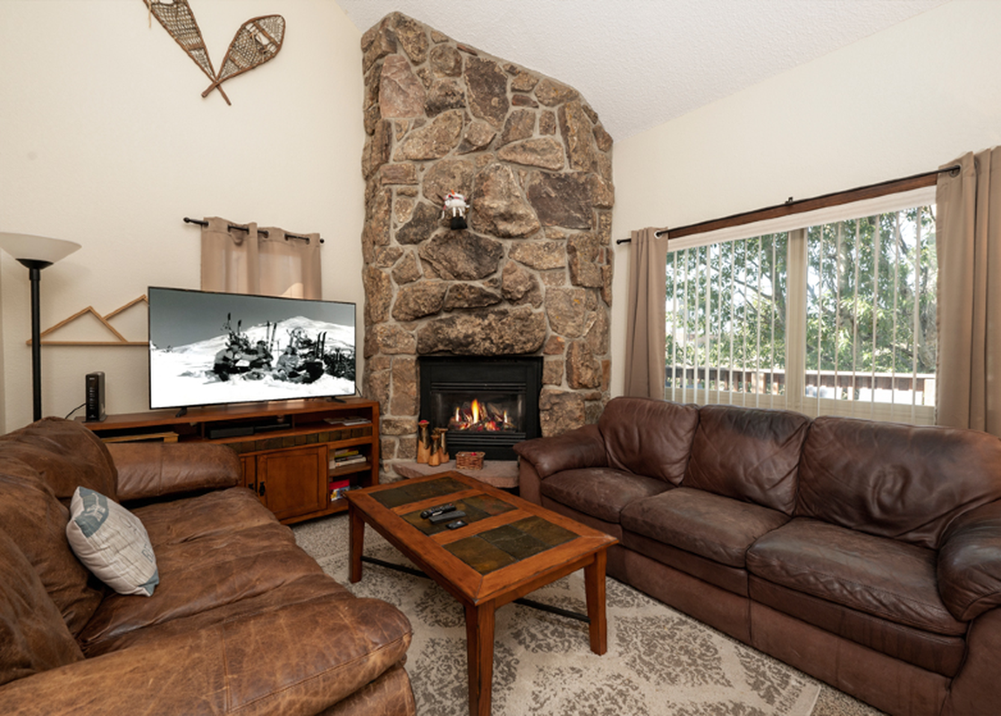 The inviting living area features plush leather sofas, a large flat-screen TV, and a cozy fireplace, all set against a backdrop of natural light from the large windows.
