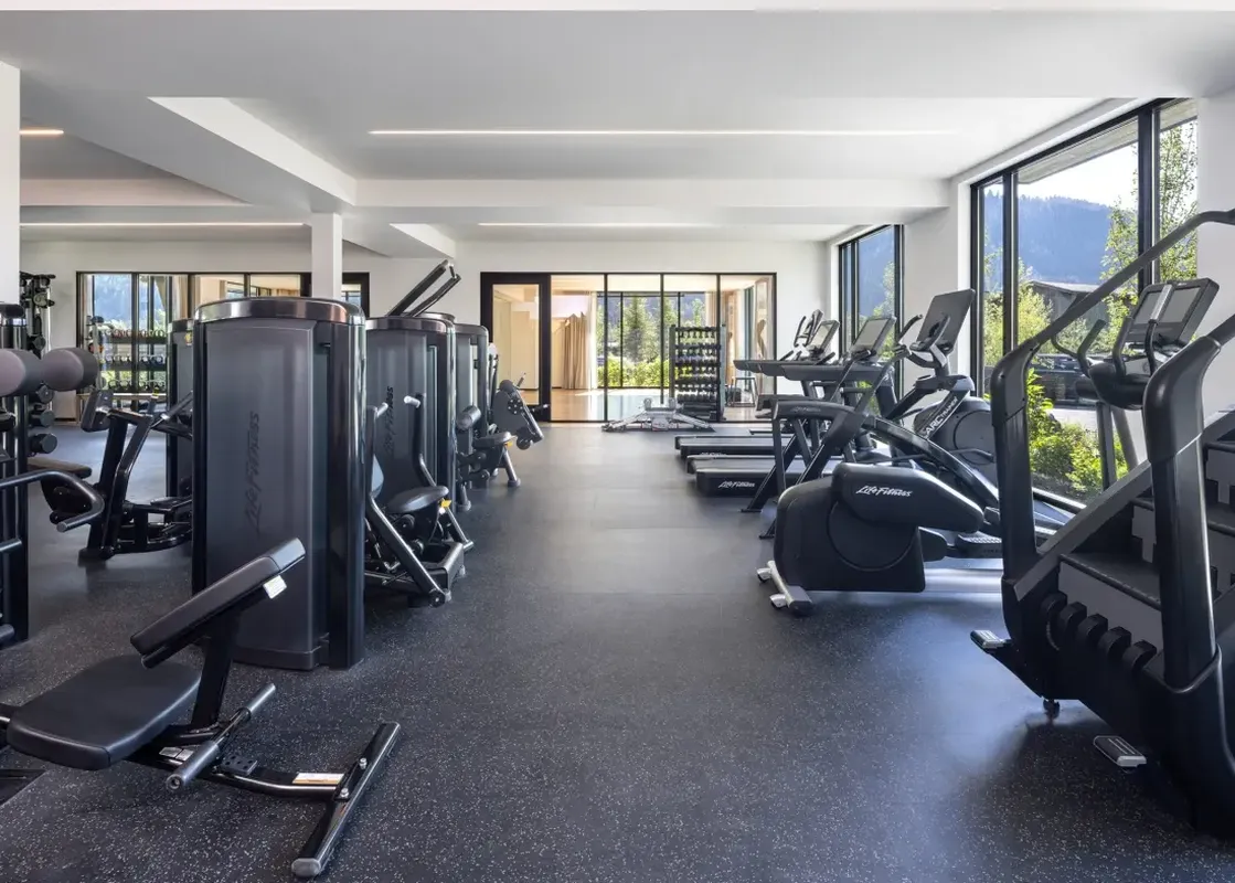 A modern and spacious fitness center with a variety of exercise machines, including treadmills, ellipticals, stair climbers, and strength training equipment, set against a backdrop of large windows offering mountain and forest views.