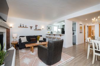 A stylish open-concept living area with a dark gray sofa, leather armchair, live-edge coffee table, and a breakfast bar leading to a modern kitchen.
