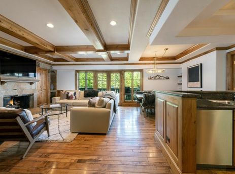A spacious open-concept living and dining area with hardwood floors, a stone fireplace, comfortable seating, and a dining table with a view of green trees outside.