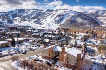 An aerial view of the snow-covered ski resort town, with ski slopes in the background, under a bright blue sky with white clouds.