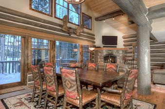 A cozy dining area in Powder Ridge Cabin Manitou 1 with a wooden table, patterned chairs, large windows overlooking a snowy forest, and a stone fireplace with a TV.