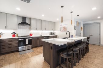 A modern kitchen in The Aerie features light gray and dark wood cabinetry, stainless steel appliances, a large island with bar stools, and elegant pendant lighting.