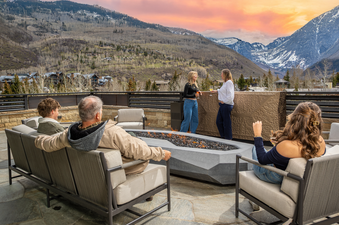 An outdoor seating area at the Opus Estate features comfortable modern sofas and chairs surrounding a fire pit, with majestic mountains and a beautiful sunset in the background.