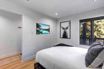 A serene bedroom at Snowcrest 216 with white walls, a large bed with white linens, and two framed art pieces on the wall, one of a mountain lake and another of a stag, complemented by a large window overlooking trees.