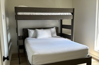 A bunk bed with a double bed on the bottom and a single bed on the top is featured in a bright room with light wood flooring and a modern ceiling fan.
