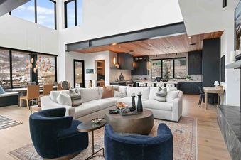 A modern, open-concept living room with a large white sectional sofa, navy armchairs, a circular coffee table, and an adjacent dining area and kitchen, all set against a backdrop of snowy mountain views through expansive windows.