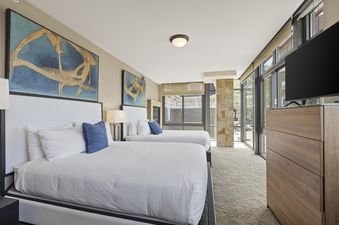 A bright bedroom in Lowell 401 with two queen beds, each featuring white linens and a blue throw pillow, under a large piece of abstract art.