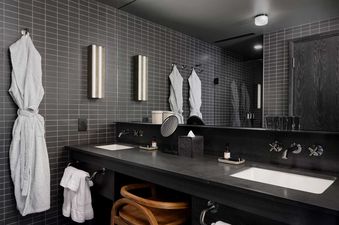A modern bathroom in the Mollie Aspen Mollie Suite features dark tiled walls, a double vanity with black countertops, and a large mirror.