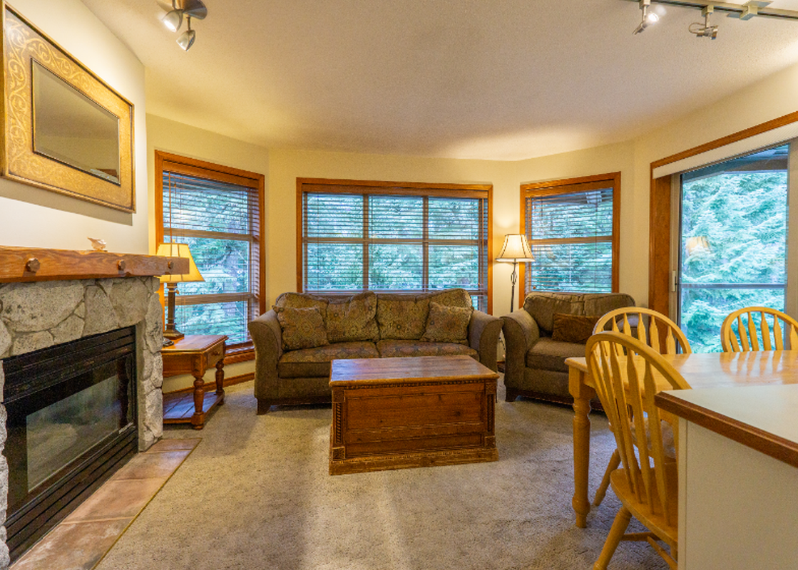 The living area of Aspens on Blackcomb Unit 427 features a comfortable sofa and armchair, a coffee table, and a stone fireplace, with large windows overlooking a lush exterior.