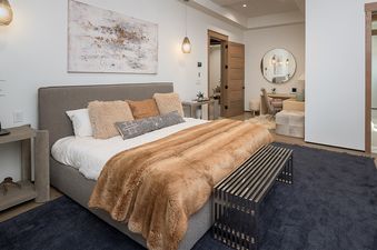 A luxurious primary bedroom featuring a king-sized bed with a faux fur throw, stylish pendant lights, and an abstract artwork, with an en suite bathroom and workspace visible through an open doorway.