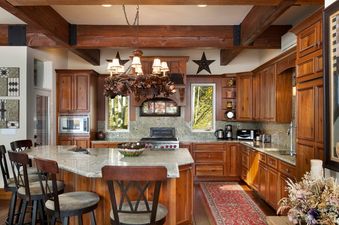 The kitchen at 185 Nighthawk Dr features rich wooden cabinetry, a large granite island with bar stools, and exposed wooden beams with a unique chandelier, complemented by modern appliances and picturesque window views.