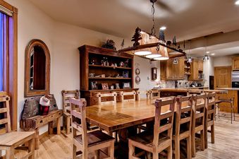 A rustic dining area with a long wooden table and chairs, a large hutch, and a kitchen area in the background at His High Place.