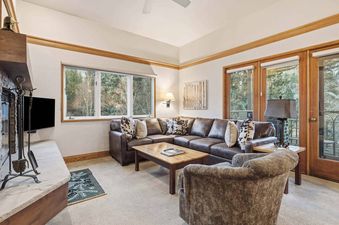 A cozy living room in The Charter E2105 features a large brown sectional sofa with cow-hide pillows, a coffee table, a stone fireplace, and a large window with an outdoor view of trees.