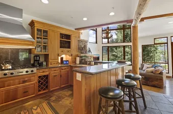 A spacious kitchen with wooden cabinetry, black granite countertops, a stainless steel range, and a breakfast bar with three stools, leading to a living area with large windows showcasing outdoor greenery.