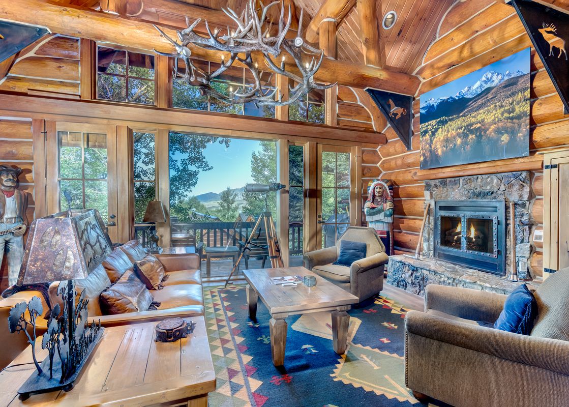 A rustic and cozy log cabin living room at Summit Cabin See Forever Cabin 103, featuring comfortable seating, a stone fireplace, and panoramic mountain views through large glass doors.