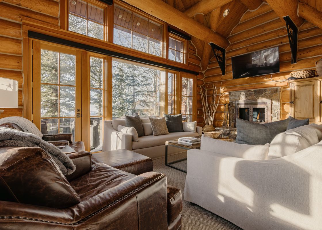 A cozy living area within Frostpine Cabin at See Forever, featuring exposed log walls, comfortable seating, and large windows with views of snowy trees.