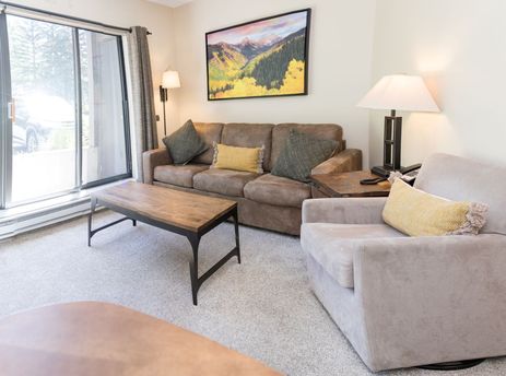 A cozy living room in the Beaver Run Resort Studio features a brown sofa, a beige armchair, and a wooden coffee table, all set on a light-colored carpet.