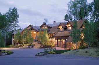 A large, multi-level chalet-style home at dusk, with warm lights glowing from its windows, nestled among tall trees, with a winding driveway and landscaped gardens leading to its entrance.
