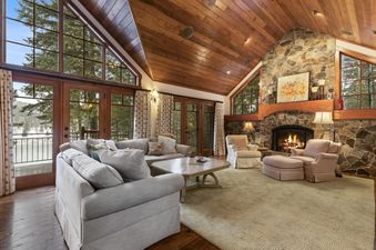 A spacious living room with a vaulted wood ceiling, stone fireplace, and large windows showcasing a snowy outdoor scene.