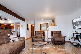 A cozy living room in Ranch at Steamboat RA101 with brown leather sofas, a cowhide rug, and a TV showing a snowy mountain scene, with a kitchen visible in the background.