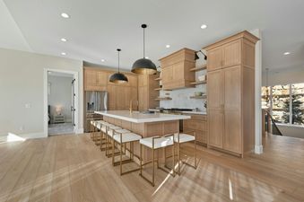 A modern kitchen showcasing light wood cabinets, a spacious island with a white countertop, two black pendant lights, and six gold-framed bar stools, offering a blend of modern and classic design.