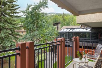 A balcony with two chairs and a small table with a vase of flowers, overlooking a lush green landscape with trees and distant mountains under a cloudy sky.