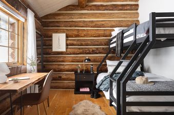 A cozy bunk bedroom at Rodeo Ridge featuring log cabin walls, a sleek black metal bunk bed, a wooden desk with a leather chair, and a soft faux fur rug on the floor.