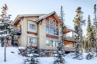 A large, multi-story luxury suite, Cowboy Heaven Luxury Suite Unit 2B, is surrounded by snow-covered pine trees under a clear winter sky, showcasing its rustic yet elegant exterior.