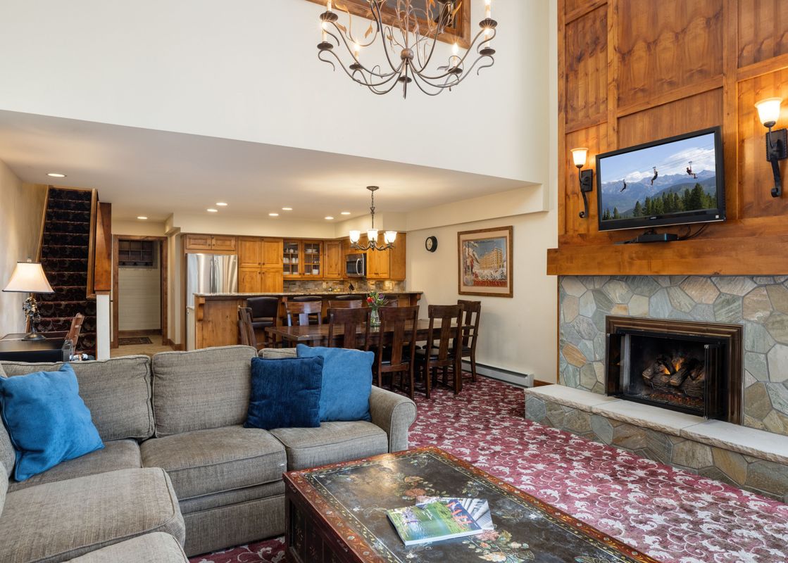 A cozy living room at Montaneros 418 featuring a sectional sofa, a stone fireplace with a TV above it, and a dining area connected to the kitchen.