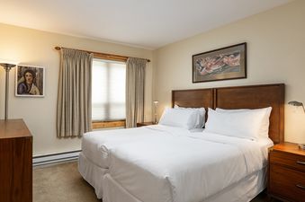 The bedroom of Montaneros 202 features two beds with white linens, wooden headboards, and bedside tables with lamps. A window with blinds and drapes is visible, along with two framed artworks on the walls.