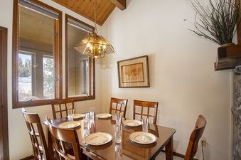 A cozy dining area features a wooden table set for six with light-colored place settings and glassware, beneath a classic chandelier, offering a view of snow-covered trees through large windows.