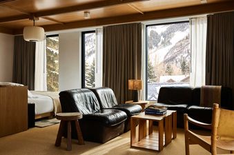A modern guest room features a wooden ceiling, two large windows with mountain views, black leather seating, and a large bed.
