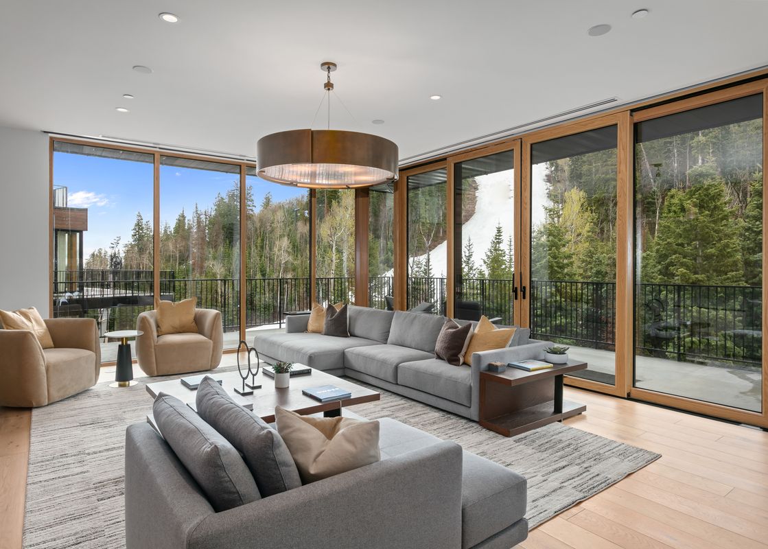 A spacious living room at Founders Place 1504 with a large gray sectional sofa, two beige armchairs, a modern coffee table, and expansive windows offering views of a forested mountain landscape.