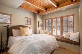 The comfortable queen-sized bed in the bedroom of Moonlight Mountain 38B Cowboy Heaven, featuring a brown leather headboard, white linens, and decorative pillows, is flanked by windows with wooden blinds revealing a snow-covered exterior.