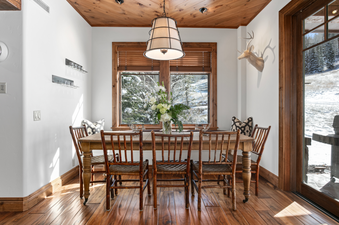 The dining area in Grand Lodge Four Bedroom 306 has a large wooden table with six chairs, a white, wooden chandelier, and a view of the snow-covered mountains.