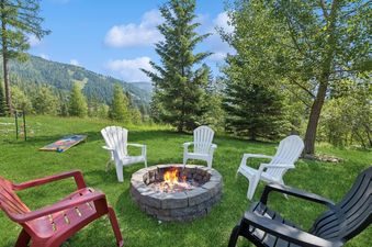 An outdoor fire pit area at the Chair View Chalet, featuring Adirondack chairs around a stone fire pit with a fire burning, set against a backdrop of lush green mountains under a blue sky.