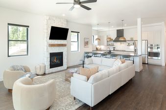 A modern open-concept living and kitchen area at Alpine Proper featuring a stone fireplace, light-colored sofas, and a sleek kitchen with golden pendant lights.