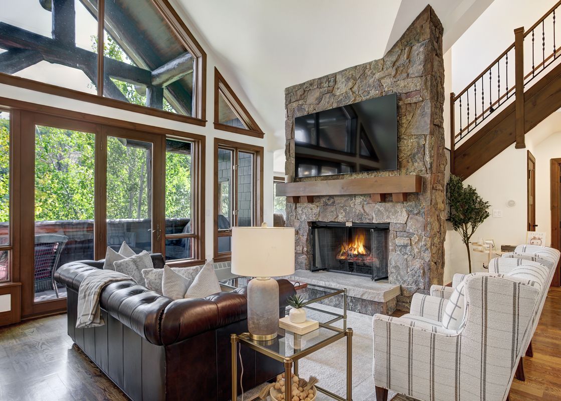 A cozy and elegant living room featuring a large stone fireplace, a comfortable leather sofa with throw pillows, and tall windows looking out onto a vibrant green landscape.