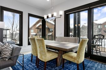 A modern dining room in Juniper 901 features a wooden table with yellow upholstered chairs and a bench, set against a backdrop of large windows offering snow-covered mountain views.