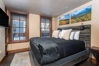 The primary bedroom features a plush king-sized bed, two large windows, a flat-screen TV, and mountain landscape artwork.